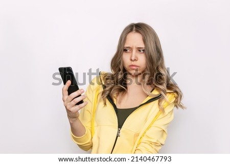 A frightened young caucasian blonde woman with wavy hair holds a mobile phone in her hand looking suspiciously at the incoming call from an unknown number isolated on a white background. Fraud
