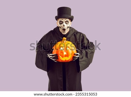 Frightening crazy man in masquerade costume frightfully exclaims frighteningly on Halloween. Emotional man in black cloak, hat and with painted skeleton on his face holding pumpkin on lilac background