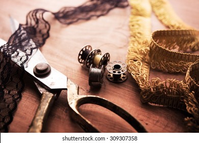 Fringe or lace tapes and silk trimmings with gold (brass) scissors and old sewing machine bobbins on a old grungy work table. Tailor's workbench. textile or fine cloth making.Shallow depth of field.