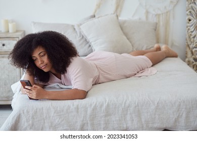 A Frizzy-haired, Good-looking Afro American Woman Lies On A Bed With A Smartphone, Spending Weekend Chatting On Social Media, Using A New Mobile App, Having Fun On The Dating App.