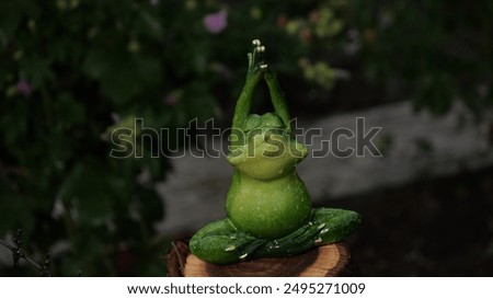 A frog statue is sitting on a log, with its hands in a prayer position. The statue is green and he is a representation of a frog