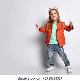 Frolic active cheerful blond baby girl kid in leather jacket, white t-shirt, blue jeans and white sneakers shows V-signs victory with both hands on background with free copy space