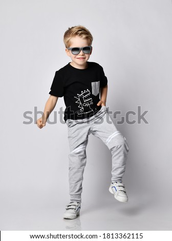 Frolic blond kid boy in sunglasses, black t-shirt with dinosaur print and gray pants stands with his foot up stamping loudly over gray background