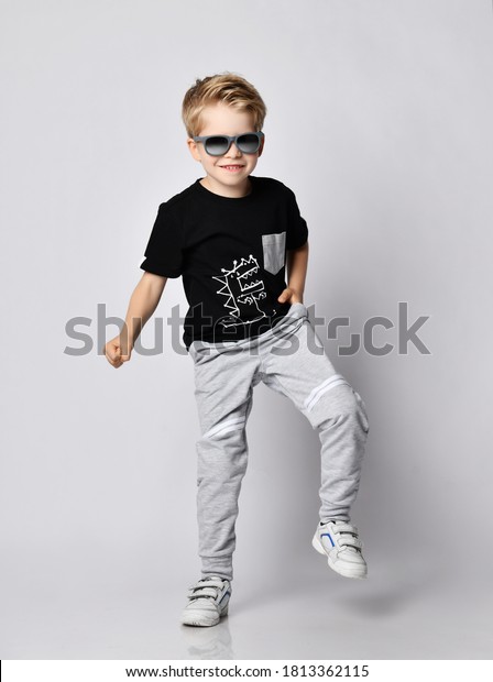 Frolic blond kid boy in sunglasses, black\
t-shirt with dinosaur print and gray pants stands with his foot up\
stamping loudly over gray\
background