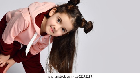 Frolic curious kid girl with straight brunette hair with buns in modern fashion pink brown sportwear is bending over to peek in camera on gray background with free text copy space