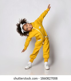 Frolic dark-skinned african american curly kid girl in yellow sport jumpsuit performs sideways bands holding arms spread wide like wings, warmup, morning gymnastics in sport school, kindergarten