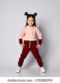 Frolic Smiling Kid Girl With Straight Brunette Hair With Buns In Two-colored Pink Brown Sport Suit Hoodie And Pants Poses With Legs Wide Apart And Hands On Hips