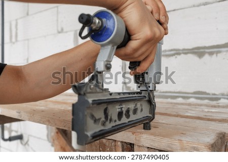 A front angle view of a pneumatic brad nailer being held by hands against a wooden surface on a construction site.