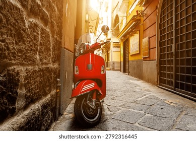Front angle view of a vibrant red motor moped stationed next to an ancient stone wall in a sunlit narrow street.