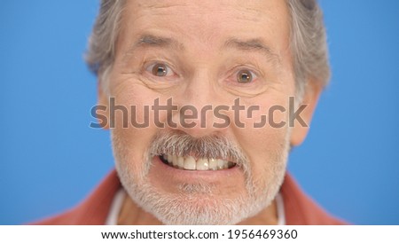 In front of the blue background, bearded old man clenches his teeth, nervous face plan.