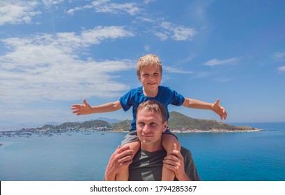 Front Boy Son Laugh Sitting On Fathers Shoulders. No Photoshop Sun On Skin. Sea, Clouds, Island Background. Funny Photo, Happiness Lifestyle, Father's Day, Love Parenthood, Family Holiday Concept