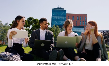 In front of the camera charismatic multiracial group of students doing a college project together using laptops and tablets to studying in the middle of modern college park