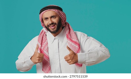 In front of camera, Middle Eastern guy gives thumbs up indicating consent and fulfillment while dressed in customary Muslim apparel, young person wearing traditional Arab clothing, like symbol.