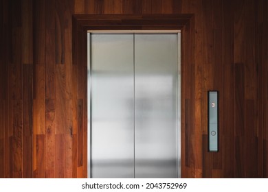 In front of the closed shiny metallic lift elevators, with a brown wooden wall background in the building
