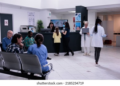 Front Desk Receptionist Talking With Young Girl Patient Registering For Doctor Appointment With Her Mother In Private Hospital. Nurse Talking With Diverse Patients Waiting In Modern Clinic.