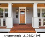 Front door and covered porch of new home exterior: solid wood door is flanked by sconce lights and has glass panels in upper portion and mullions.