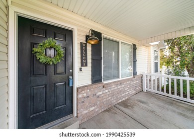 Front door exterior with leaf wreath, wooden white railings and wall bricks. There is a door ringer and decorative light and display beside the door and a window with fake shutter design.