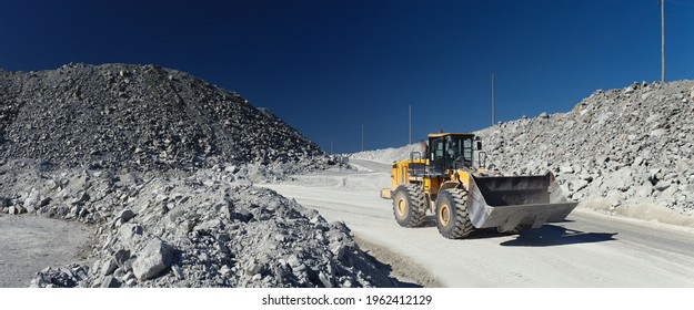 Front end loader excavator moves along the road in a stone quarry against the background of large stone dumps and a dark blue sky, panorama.