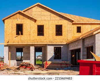 Front exterior of single-family house under construction, with stacks of lumber and a dumpster (bottom right) near two-car garage, in a suburban development on a sunny morning in southwest Florida