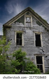 The Front Facade Of An Abandoned Haunted House With Blown Out Windows, Bare Rotting Wood And Ugly Overgrown Shrubbery