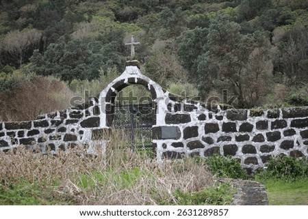 A front facade of the old stone cemetery wall with an arched entrance and cross, leading to a wrought-iron gate in the Village Faja of Caldeira de Santo Cristo saint Jorge island azores 