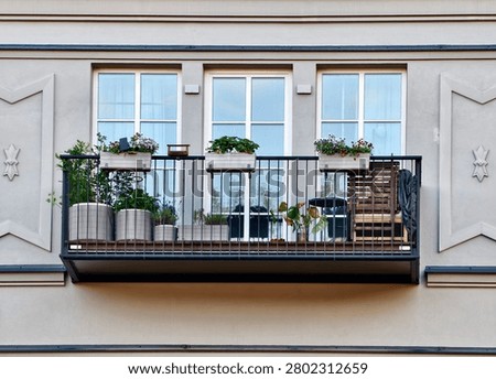 Front facing view of compact apartment balcony featuring metal railing, three planter boxes with blooming flowers, several large pots with greenery, white framed windows, and pale stucco facade