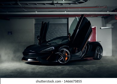 Front left quarter of black Mclaren 570s with orange accents and doors open, featuring smoke in background.