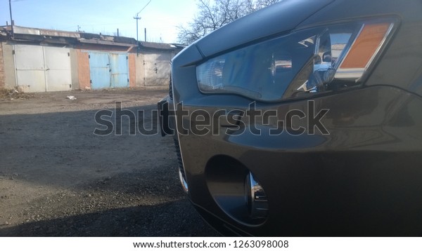 Front left side and headlight of a modern\
brown car with a lower left fog lamp against the background of old\
Soviet garages on an autumn afternoon. Texture of the Japanese\
expensive car crossover.