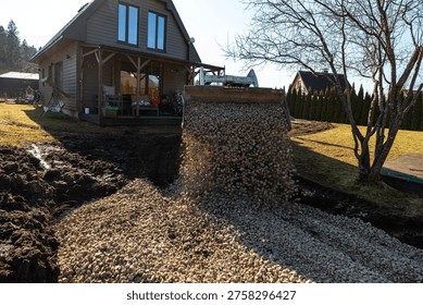 A front loader tips crushed stone into a dug path by a timber clad house with porch. Pebbles fly mid air on a sunny day, curving route past a bare tree and hedges.