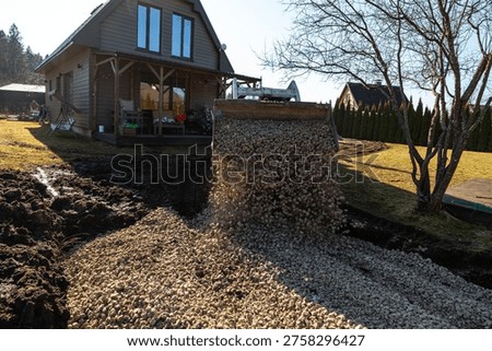 A front loader tips crushed stone into a dug path by a timber clad house with porch. Pebbles fly mid air on a sunny day, curving route past a bare tree and hedges.