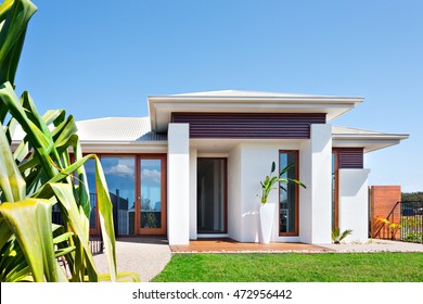 Front Of The Modern Villa With A Lawn And Blue Sky, The House Has A Small Green Grass Of The Ground And The Concrete Yard Front Of It. The Facade Has A Tall And White Vase With An Ornamental Plant. 