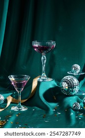 A front perspective of a raised emerald surface with cocktail glasses with purple water, confetti, golden ribbon, and a few tiny disco balls set against a green backdrop. A template for a party theme.