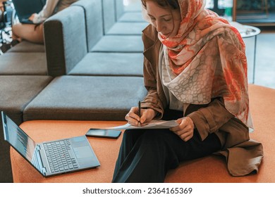 Front photo of muslim businesswoman, searching information, analyzing data, sales reports and profit growth while working at the office sitting on an orange sofa - Shutterstock ID 2364165369