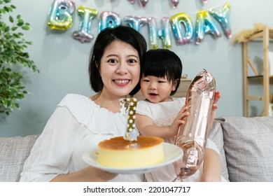 Front Portrait Of A Curious Asian Baby Girl Looking At The Birthday Cake While Her Mother Is Smiling At The Camera In A Party At Home