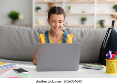 Front portrait of smiling female teenager sitting on couch at desk using laptop computer at home. Happy youth looking at screen watching movie, doing homework or making video call with teacher