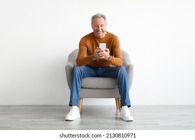 Front Portrait Of Smiling Mature Man Using Mobile Phone, Watching Video Sitting On Armchair Isolated On White Studio Background Wall. Guy Browsing Internet, Surfing Web, Free Copy Space