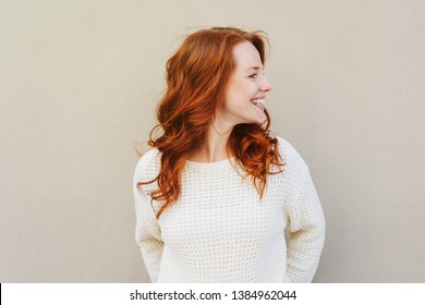 Front portrait of young red-hared woman in white sweater, smiling and looking away, standing against grey wall with her head turn left