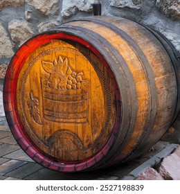 In front of a rustic stone wall lies a wooden barrel, with its top surface adorned by wood carvings of grape clusters, vine leaves, and a wooden tub motif, beautifully lit by the afternoon sun. - Powered by Shutterstock