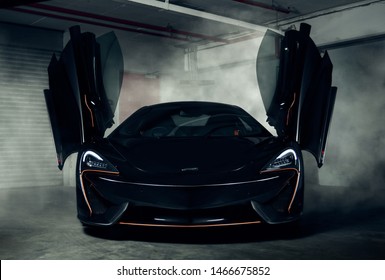 Front section of a black Mclaren 570s with orange accents and doors open, smoke in background.