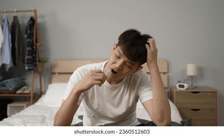 front shot of a drowsy asian young man awaking from sleep and scratching head while sitting at bedside cleaning mouth with toothbrush in the morning at home - Powered by Shutterstock