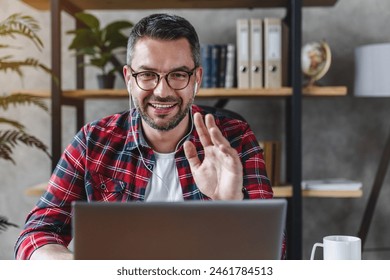 Front shot of smiling businessman freelancer entrepreneur sitting a home office looking a laptop screen having online meeting work discussion conference video zoom call, waves his hand and says hi - Powered by Shutterstock