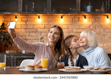 Front shot of smiling young woman taking photo selfie of her kissing grandmother daughter while having breakfast on modern kitchen. Friendly close family doing self shoot for memories - Powered by Shutterstock