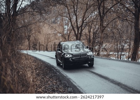 Front side view of a black modern German SUV driving along an asphalt road through an autumn or early spring landscape.