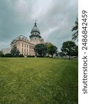 Front side view of the Illinois State Capitol Building in Springfield, IL, USA. Cloudy overcast skies overhead. Springtime scene with a lush green lawn and full trees.