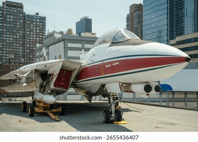 Front and side view of a white and red Navy fighter jet with visible cockpit, missiles, and landing gear. The aircraft is parked on a museum deck surrounded by city buildings.