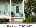 Front steps and porch of a mint green house with landscaping and potted plants. White front door with an American flag. Pumpkin decor on porch. 