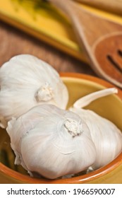 Front, Top View, Medium Distance Of Three Garlic Heads In A Yellow, Clay Ceramic Bowl With Slatted Wood Spoon