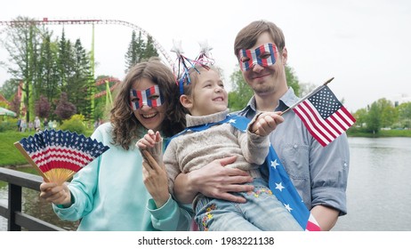 Front View. 4th July Celebration. Portrait Of Happy Young Family Standing Outside In A Park During Independence Day, Looking At The Camera, Waving Usa Flags.