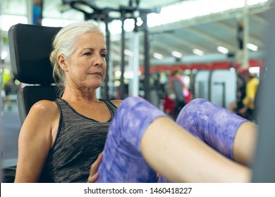 Front View Of Active Senior Caucasian Woman Exercising With Leg Press Machine In Fitness Studio. Bright Modern Gym With Fit Healthy People Working Out And Training