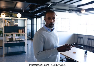 Front View Of An African American Businessman Working In The Modern Office, Standing By A Desk, Using His Smartphone And Looking Straight Into A Camera. Social Distancing And Self Isolation In